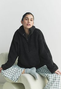 Zwarte fleece pullover met een hoge kraag en een halve rits. Draagt over lichtblauwe geruite pyjamabroek en grijze sokken. Gelaagd, warm materiaal.