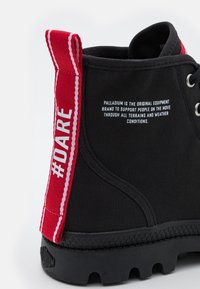 Bottine noire à la cheville avec languette de tirage rouge marquée "#DARE", texte blanc sur le côté indiquant que la marque soutient le mouvement à travers tous les terrains et conditions météorologiques.
