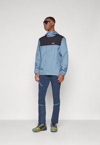 Light blue waterproof jacket with black upper panel, elastic cuffs, paired with navy pants featuring blue accents. Model wears sporty sunglasses.