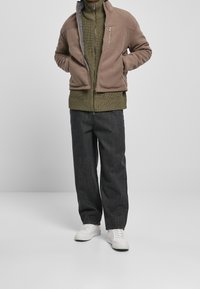 Giacca in pile marrone con colletto alto, tasca con zip, maglione a coste verde e jeans scuri larghi, abbinati a sneakers bianche.