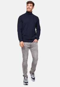 Marineblå høyhalset genser med ribbestruktur og sikksakkmønster, kombinert med lysegrå jeans og grå joggesko.