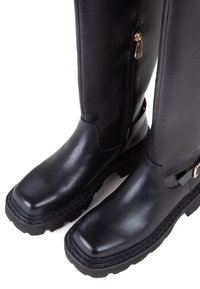 Bottes noires montantes en cuir synthétique lisse. Elles présentent une semelle épaisse, des éléments décoratifs en or et une fermeture éclair sur le côté pour un enfilage facile.