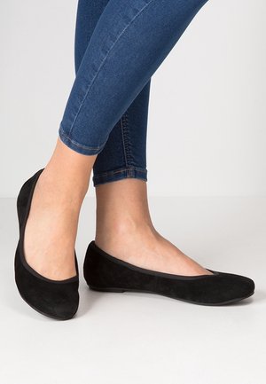 Frau trägt schwarze Ballerinas aus Wildleder mit abgerundeten Zehen, kombiniert mit dunkelblauen Skinny Jeans vor einem schlichten hellen Hintergrund.