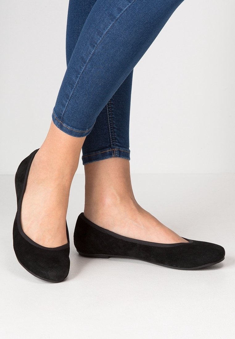 Femme portant des ballerines en daim noir à bouts arrondis, associées à un jean skinny bleu foncé, sur un fond clair uni.