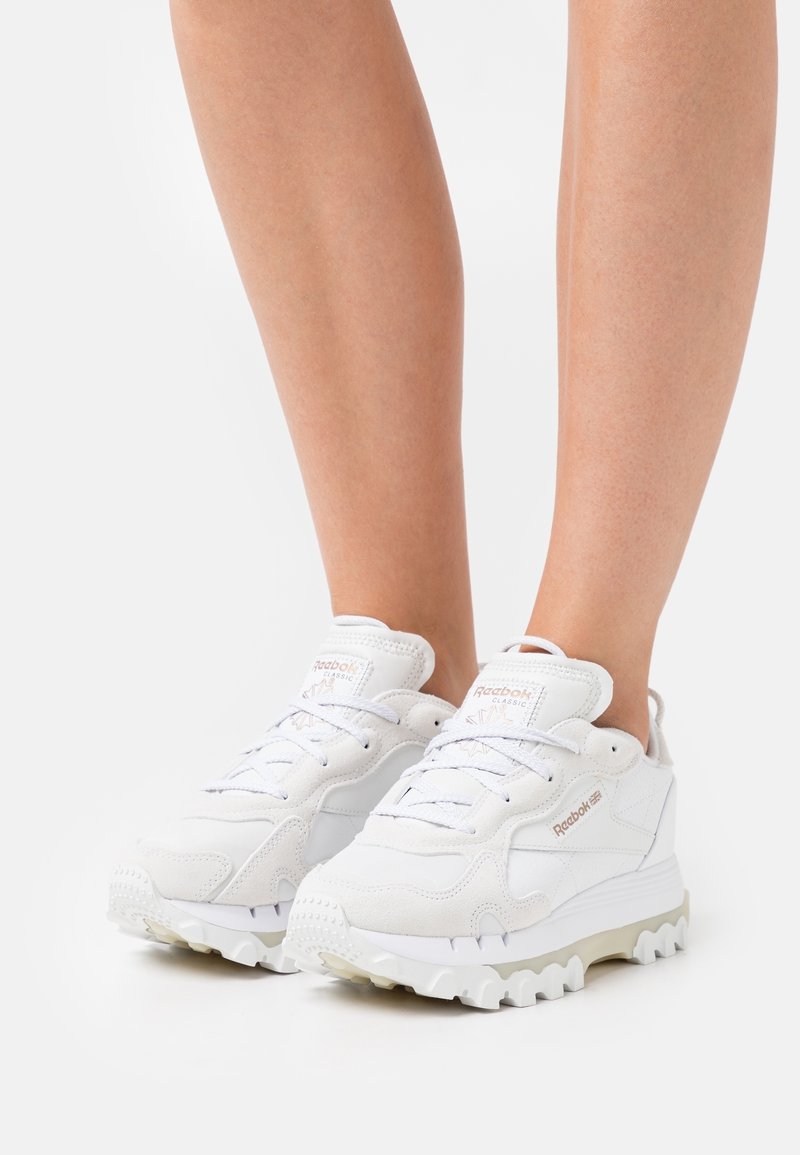 Kraftiga vita Reebok Classic sneakers med tjocka, texturerade sulor som bärs på nakna ben mot en ren vit bakgrund.