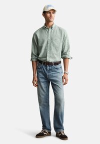 Hellgrünes Hemd zum Knöpfen, mittelblaue Jeans, brauner Gürtel und braune/weiße Turnschuhe. Beinhaltet eine beigefarbene Mütze mit hellblauem Text.