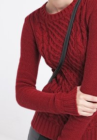 Röd stickad tröja med kabelmönster, rund hals och långa ärmar. Har ribbade ärmslut och nederkant, i kombination med en tunn svart axelväska.