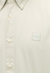 Chemise beige clair boutonnée avec un col pointu et un petit patch carré marqué "BOSS" sur la poitrine.