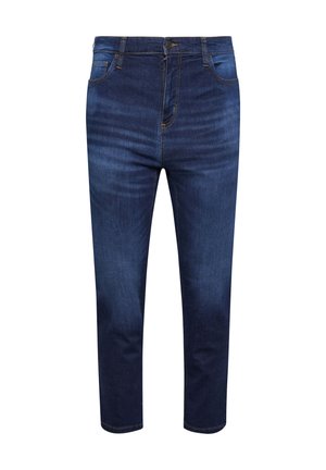Person trägt ein dunkles Shirt und blaue Jeans, steht mit einer Hand in der vorderen Jeanstasche. Nur Oberkörper und Beine sind sichtbar.