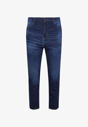 Person trägt ein dunkles Shirt und blaue Jeans, steht mit einer Hand in der vorderen Jeanstasche. Nur Oberkörper und Beine sind sichtbar.