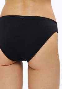 Czarne teksturowane majtki kąpielowe noszone przez osobę, ukazujące dolną część pleców i górną część ud, z małą metką marki umieszczoną na środku pasa.