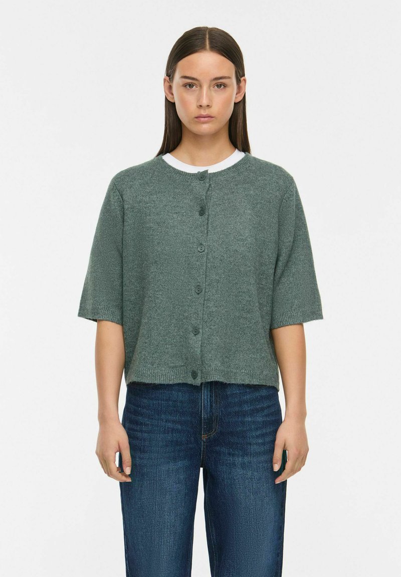 Femme aux cheveux raides et foncés porte un cardigan vert à manches courtes boutonné sur un haut blanc associé à un jean bleu, debout devant un fond blanc.