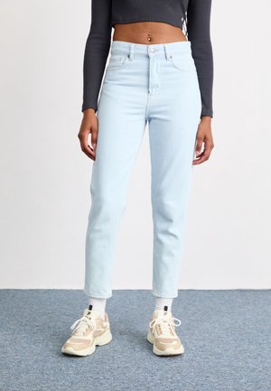 Persoon die lichtblauwe high-waist jeans, beige sneakers met witte veters, witte sokken en een zwart cropped top met lange mouwen draagt, staand binnen.