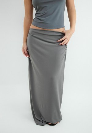 Femme portant un haut gris sans manches et une longue jupe grise, se tenant devant un fond clair uni, tenant la taille de la jupe.