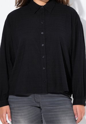 Femme portant une chemise noire ample boutonnée à manches longues et un jean gris délavé, debout devant un fond uni.