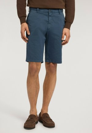 Mand iført marineblå knælange shorts, brun langærmet top og brune loafers, stående mod ensfarvet baggrund.