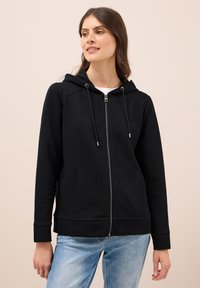 Donna che indossa una felpa nera con zip, con cappuccio e tasche frontali, abbinata a jeans azzurri chiari, che guarda leggermente di lato.