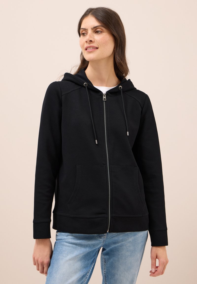 Donna che indossa una felpa nera con zip, con cappuccio e tasche frontali, abbinata a jeans azzurri chiari, che guarda leggermente di lato.