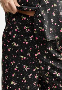 Černá látka s květinovým vzorem, který obsahuje růžové jahody a bílé květiny, texturovaná malými tečkami, zapínaná na bílé knoflíky.