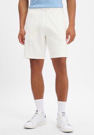 Mann trägt weiße knielange Shorts, weiße Crew-Socken und weiße Sneaker und steht vor weißem Hintergrund.