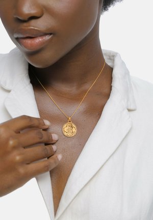 Goldene Anhängerkette mit einem runden Charm, der mit filigranen Details versehen ist, getragen um einen nackten Hals, sichtbar mit einem hellen Hemd und einer Hand.