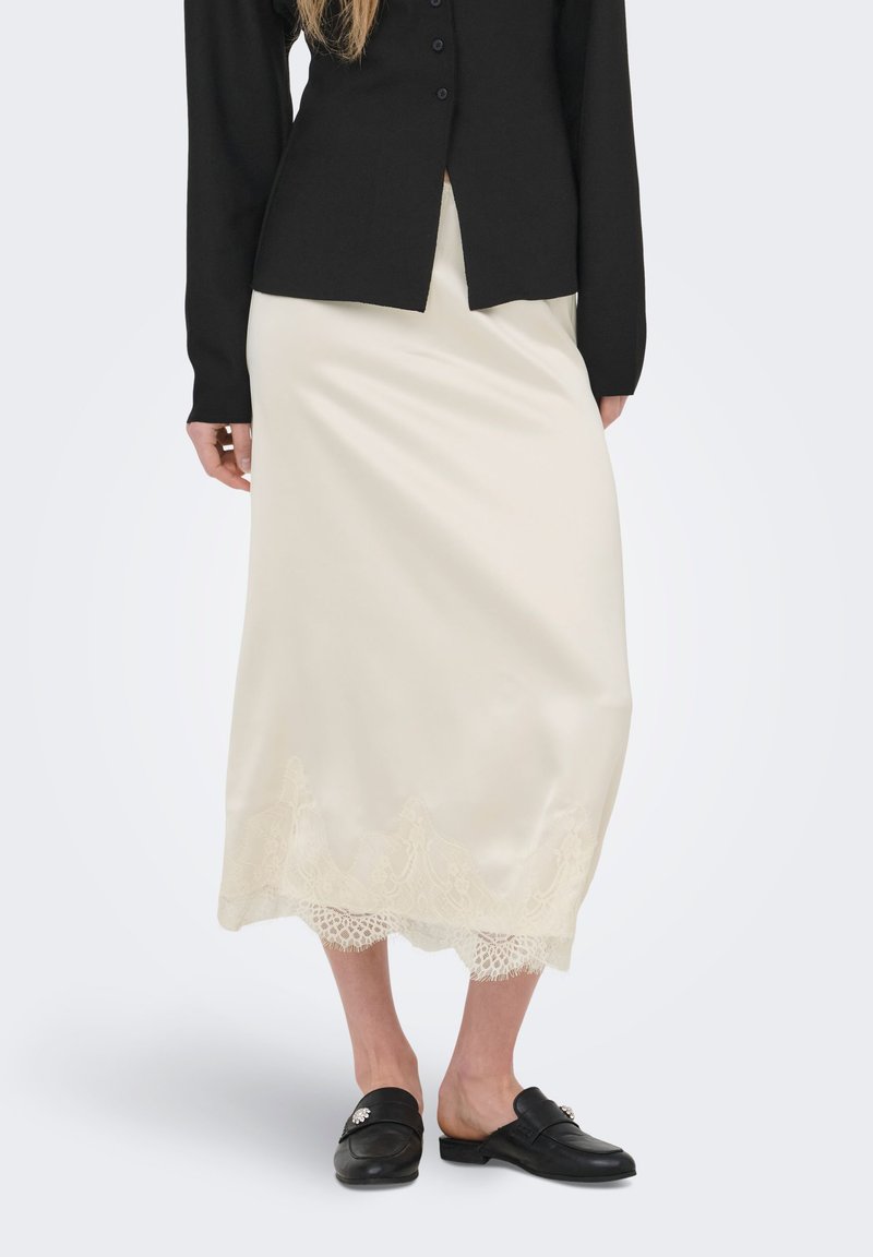 Femme portant une jupe en satin crème longueur genou avec un bord en dentelle, une veste noire boutonnée et des chaussures noires sans lacets avec des accents décoratifs.