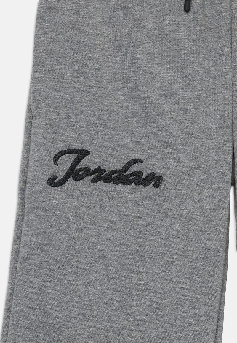 Pantalon de survêtement gris en tissu doux, présentant un grand logo "Jordan" noir brodé sur la cuisse et une taille avec cordon de serrage.