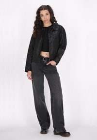 Zwarte cropped jas met camouflagepatroon, gecombineerd met hoge zwarte jeans. Model staat met handen in de zakken en draagt zwarte laarzen.