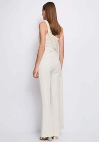 Tuta beige con design monospalla, dettaglio drappeggiato sulla schiena, corpetto aderente e gambe svasate, realizzata in tessuto liscio ed elasticizzato.