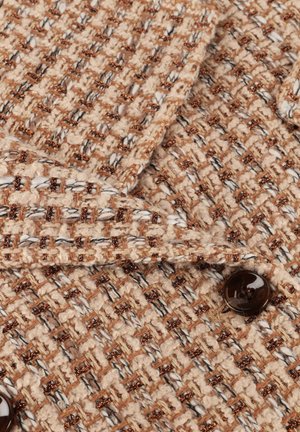 Geweven stof in beige en bruin met metalen draadaccenten. Heeft een gestructureerde oppervlakte en een donkerbruine knoopdetail.