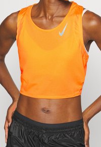 Top court Nike orange vif avec une finition texturée, encolure arrondie et détail de logo réfléchissant. Porté avec un short noir.