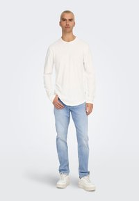 Camisa blanca de manga larga con dobladillo redondeado, combinada con jeans de un azul claro y zapatillas blancas gruesas, con un fondo claro.