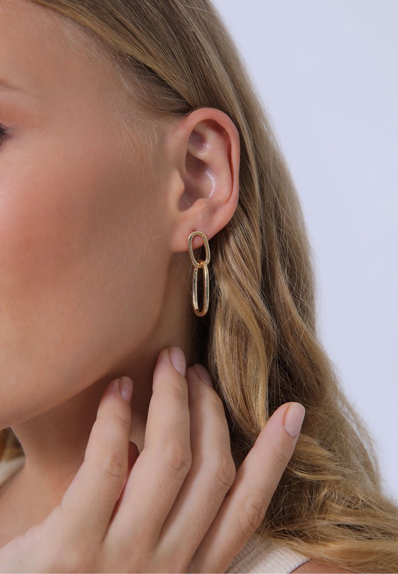 Gros plan sur l'oreille d'une femme avec deux boucles d'oreilles ovales en or liées et sa main touchant doucement son cou près de la boucle d'oreille.