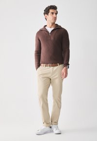 Pull en maille brun à col zippé, pantalon beige, baskets blanches, ceinture en cuir marron. Le mannequin se tient avec les mains dans les poches devant un fond uni.