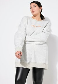 Felpa grigia chiara con la scritta "COOL & KIND" in arancione. Presenta una vestibilità comoda, polsini a coste e un'orlo arrotondato. Abbinata a leggings neri.