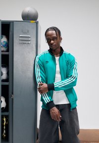 Teal Adidas treningsjakke med tre hvite striper, svarte mansjetter og logo, kombinert med grå bukser, ved siden av et grått skap med sportsballer.