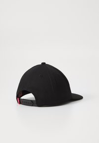 Casquette de baseball noire avec un dessus arrondi, une visière plate et une fermeture à pression ajustable. Elle présente une petite étiquette de marque rouge à l'arrière.