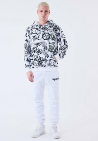 Sudadera gráfica en blanco y negro con varios estampados llamativos, combinada con pantalones de chándal blancos lisos y zapatillas blancas. Fondo simple.