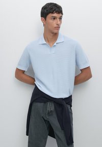 Polo shirt azzurro chiaro con un motivo a trama, maniche corte e colletto. Un maglione blu scuro è annodato intorno alla vita. I jeans grigi sono indossati sotto.