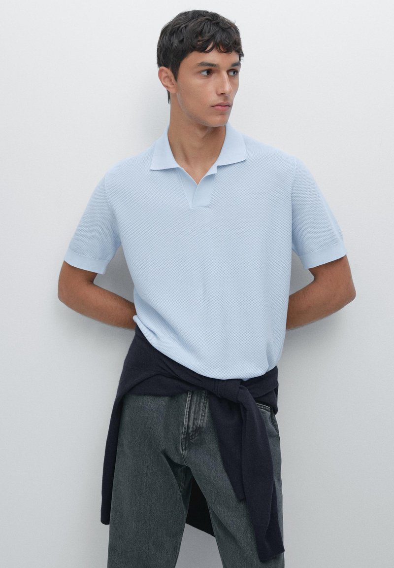 Polo shirt azzurro chiaro con un motivo a trama, maniche corte e colletto. Un maglione blu scuro è annodato intorno alla vita. I jeans grigi sono indossati sotto.