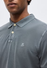 Άνδρας που φοράει γκρι πόλο πουκάμισο με δύο μπεζ κουμπιά και μικρό λευκό κεντημένο λογότυπο "MOP" στο αριστερό στήθος.