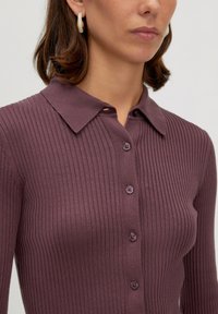 Geribbelde, lange mouw top in donker mauve met een kraag, voorzien van vijf bijpassende knopen. Vriendelijke textuur met subtiele rek.