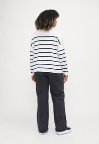 Pulover alb cu dungi bleumarin, mâneci lungi și guler rotund, purtat cu pantaloni largi gri închis și adidași albi.