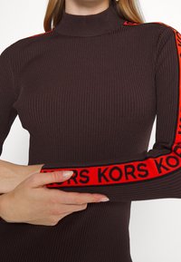 Femme portant un pull à col roulé côtelé marron avec des rayures rouges et le texte noir "KORS" le long des manches, les bras croisés sur la poitrine.