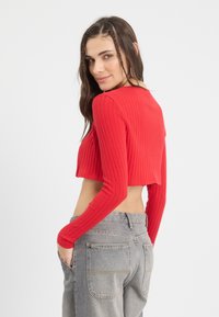 Červený pruhovaný crop top s dlhými rukávmi, má rovný výstrih a skrátený lem. Kombinovaný s šedými džínsami s vysokým pásom.