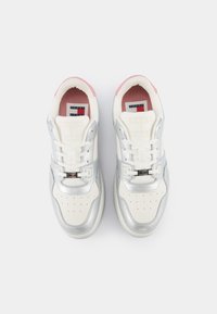 White and silver athletic shoes with a smooth texture, featuring rounded edges, laces, and a pink accent at the heel. Brand logo visible.