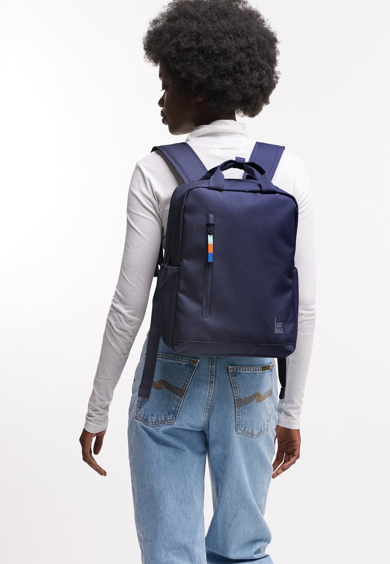 Donna con capelli ricci che indossa una camicia bianca a maniche lunghe e jeans blu che porta uno zaino rettangolare blu navy con cursore colorato.