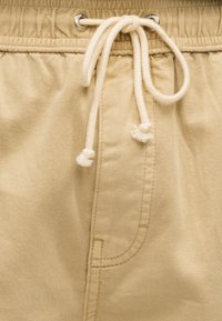 Κοντινή λήψη μπεζ βαμβακερού παντελονιού με λευκό κορδόνι δεμένο μέσα από μεταλλικά περικάρπια στη ελαστική ζώνη μέσης.