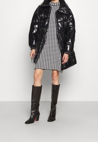 Manteau en duvet noir avec une finition brillante, taille cintrée, associé à une robe à motif pied-de-poule. Bottes en cuir noir hauteur genou avec talons bloc.