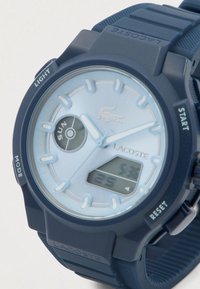 Montre-bracelet Lacoste bleue avec aiguilles analogiques, affichage numérique, indicateur de jour et boutons étiquetés lumière, démarrer, réinitialiser et mode.